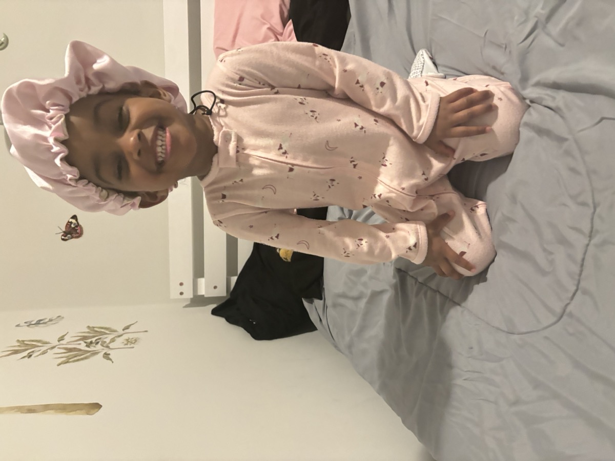 Yohanna sitting on her bed in pink pajamas and a satin bonnet, beaming with a huge smile