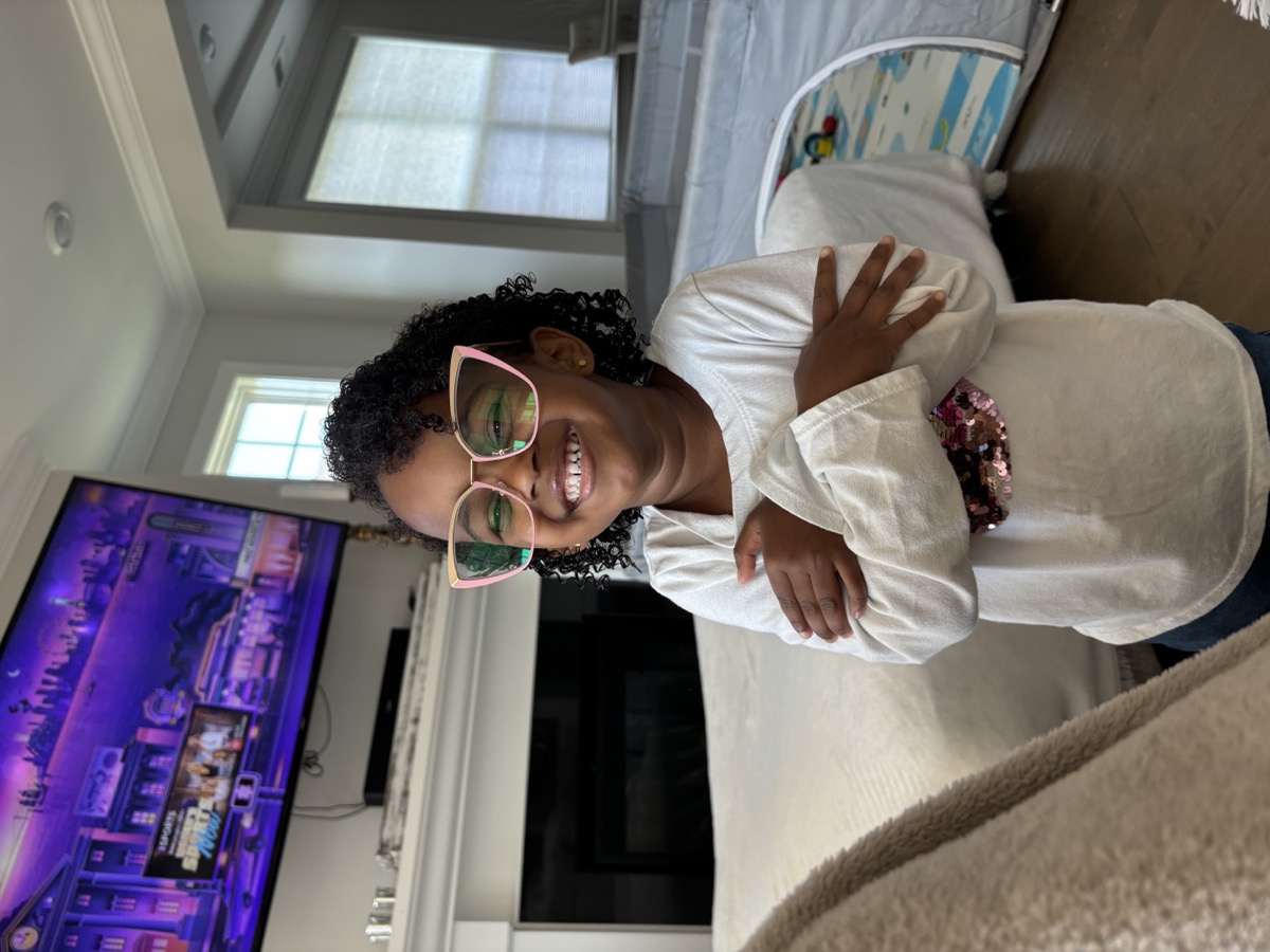 Yohanna wearing oversized pink cat-eye glasses, arms crossed and grinning confidently