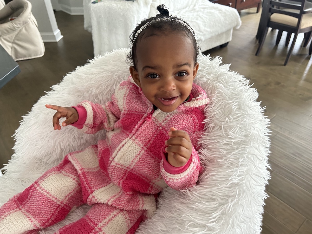 Baby Yohanna in a cozy pink plaid onesie, nestled in a fluffy white blanket and smiling at the camera
