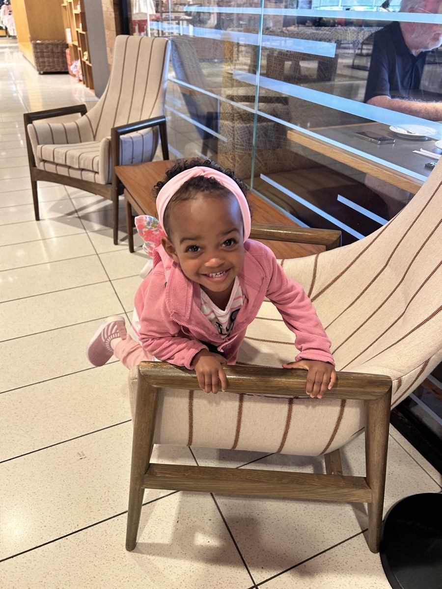 Toddler Yohanna in a pink hoodie and headband, climbing a chair with a mischievous smile