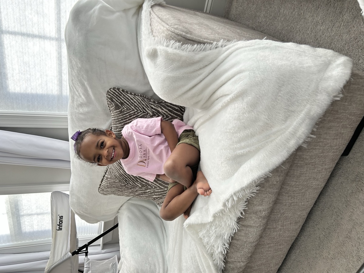 Yohanna sitting cross-legged on the couch in a pink Diosa Ara t-shirt, smiling sweetly