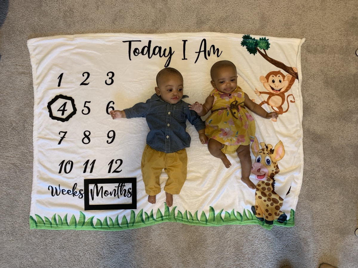 Four-month-old twins on a milestone blanket — Yohanna in a yellow floral dress and her brother in a denim shirt, holding hands