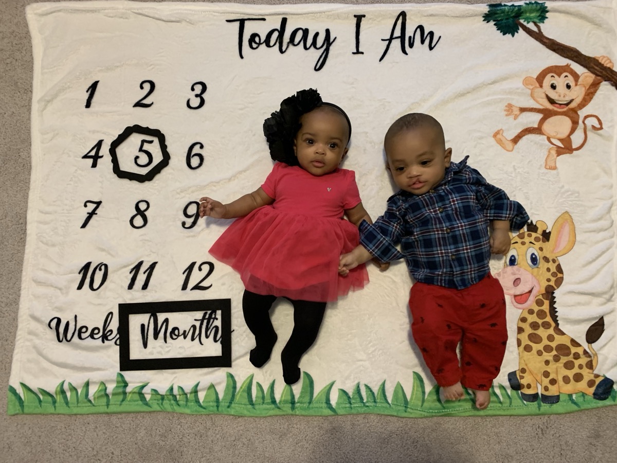 Five-month-old twins on a milestone blanket — Yohanna in a coral tutu dress with a black bow headband, holding her brother's hand