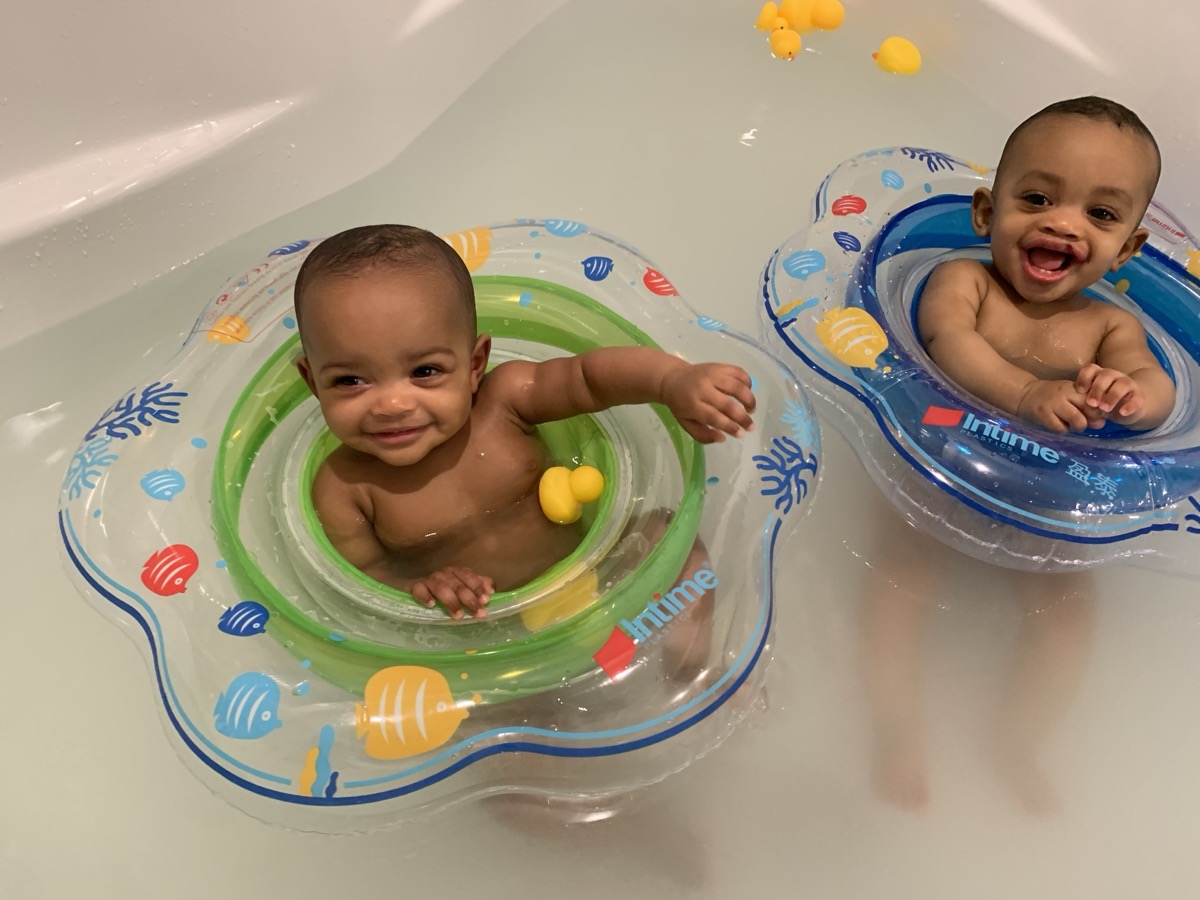 Baby twins splashing in the bathtub in inflatable float rings with rubber ducks, both grinning