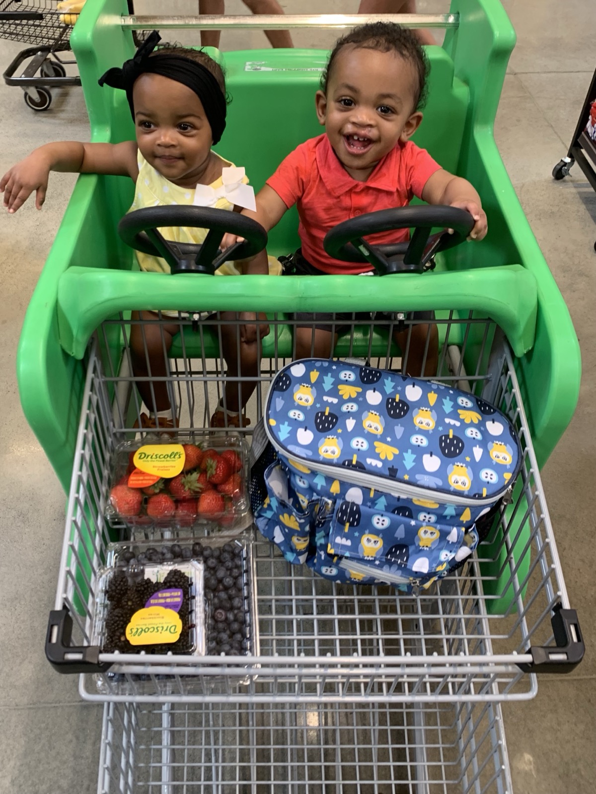 Toddler Yohanna in a yellow dress with a black headband and her twin brother in a red polo, riding in a green grocery cart with strawberries and blueberries