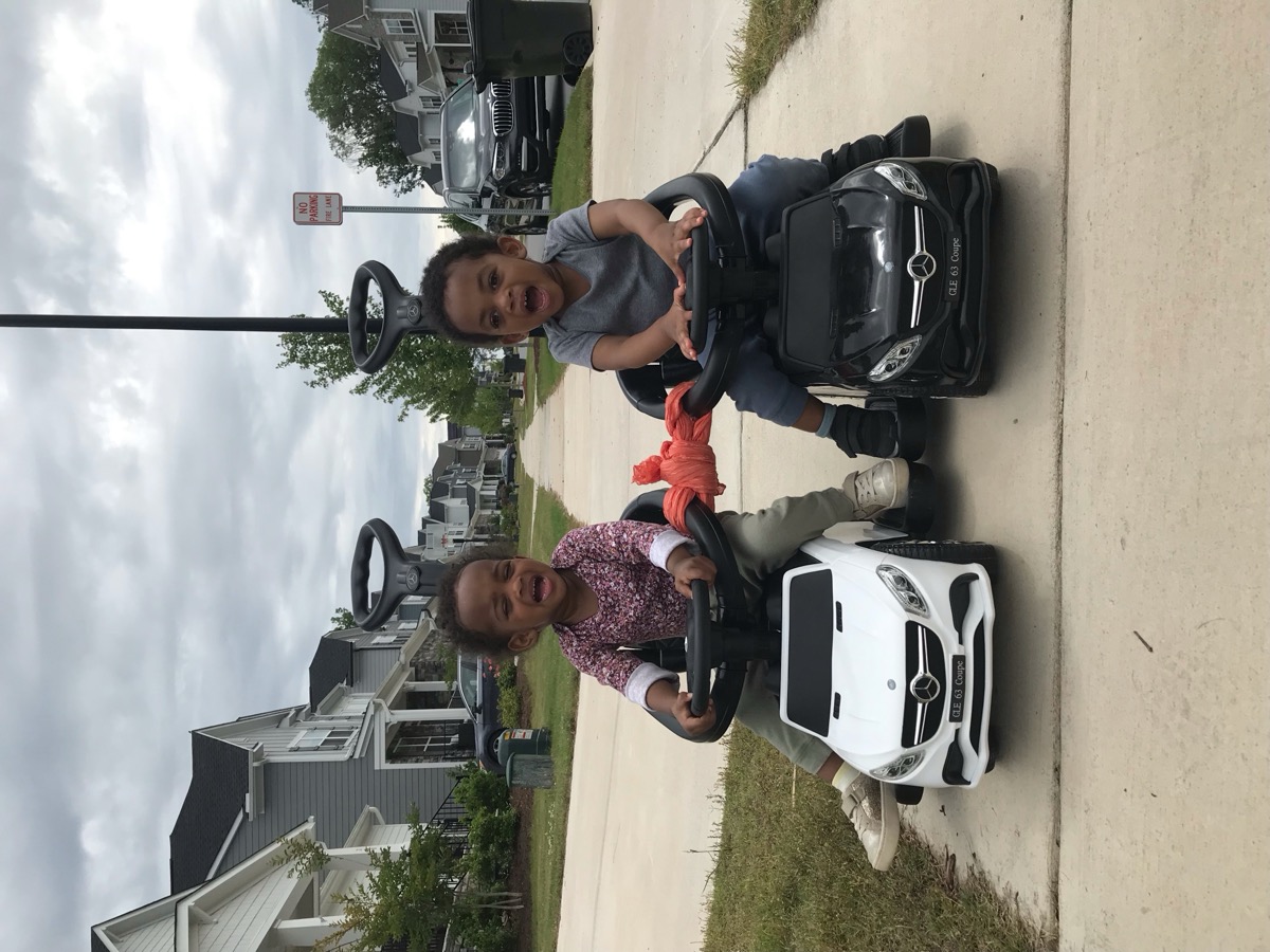 Toddler twins riding side-by-side Mercedes push cars on the sidewalk — Yohanna in a floral top on the white car, her brother in gray on the black car
