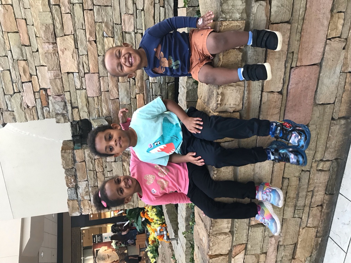 Yohanna in a pink butterfly t-shirt sitting by a stone fountain at the mall with her older brother in a shark shirt and younger brother in a bear shirt