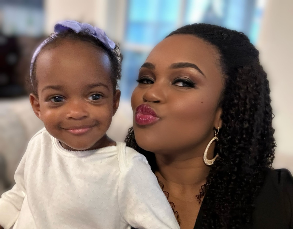 Toddler Yohanna with a purple bow headband and white sweater posing cheek-to-cheek with mom who is blowing a kiss