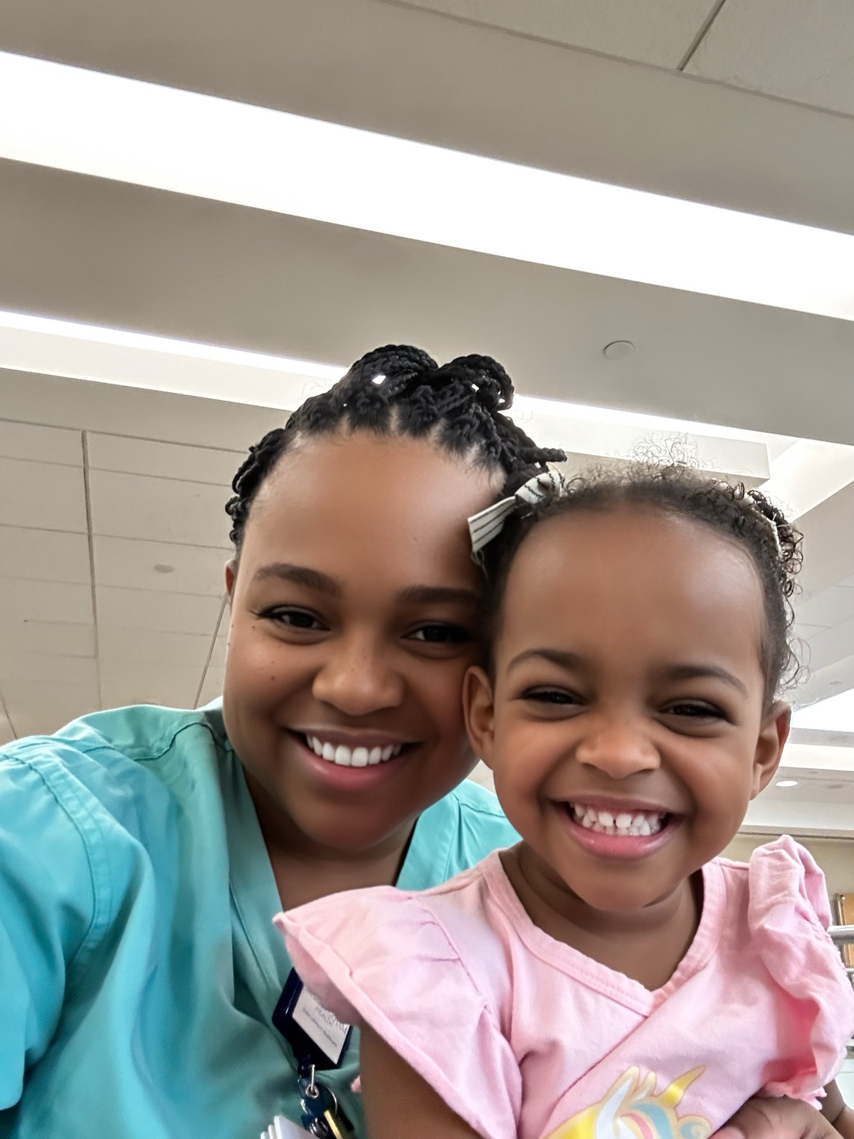 Yohanna and mom in scrubs taking a smiling selfie — Yohanna in a pink ruffled top with a striped bow in her hair