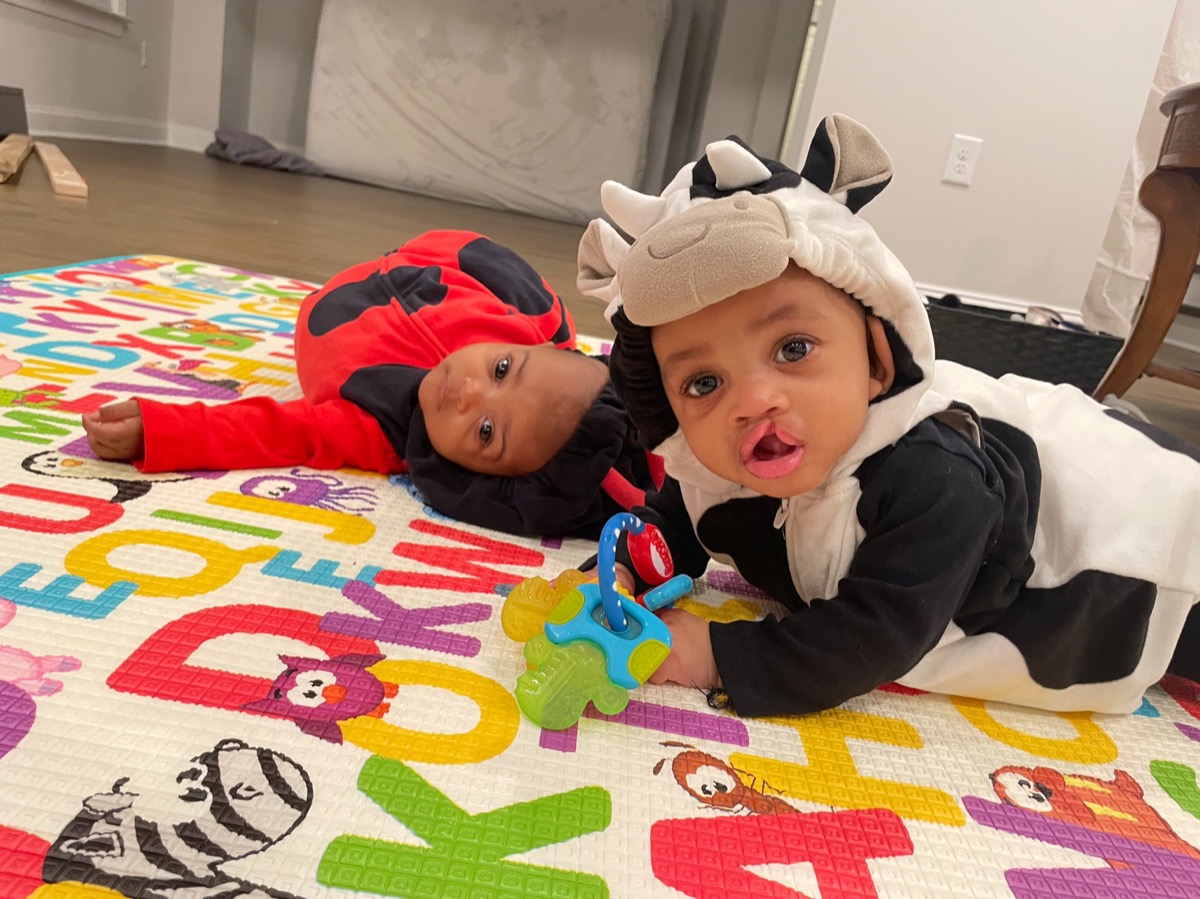 Baby twins in Halloween costumes on an alphabet play mat — one in a ladybug outfit and Yohanna in a cow costume with a teething toy