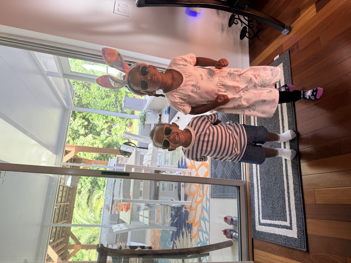 Yohanna in a pink printed dress with bunny ears headband and sunglasses, holding hands with her twin brother in a striped shirt and sunglasses by the patio door