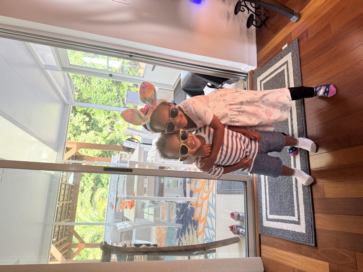 Yohanna in bunny ears and sunglasses hugging her twin brother by the patio sliding door, both smiling