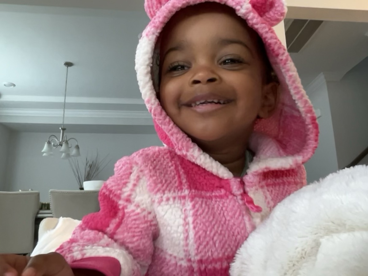 Toddler Yohanna in a cozy pink plaid bear-ear hoodie, grinning big with a fluffy white blanket