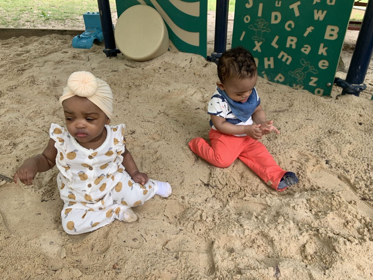 Baby Yohanna in a cream turban headwrap and a polka-dot romper sitting in the sandbox next to her twin brother in a blue bib and red pants