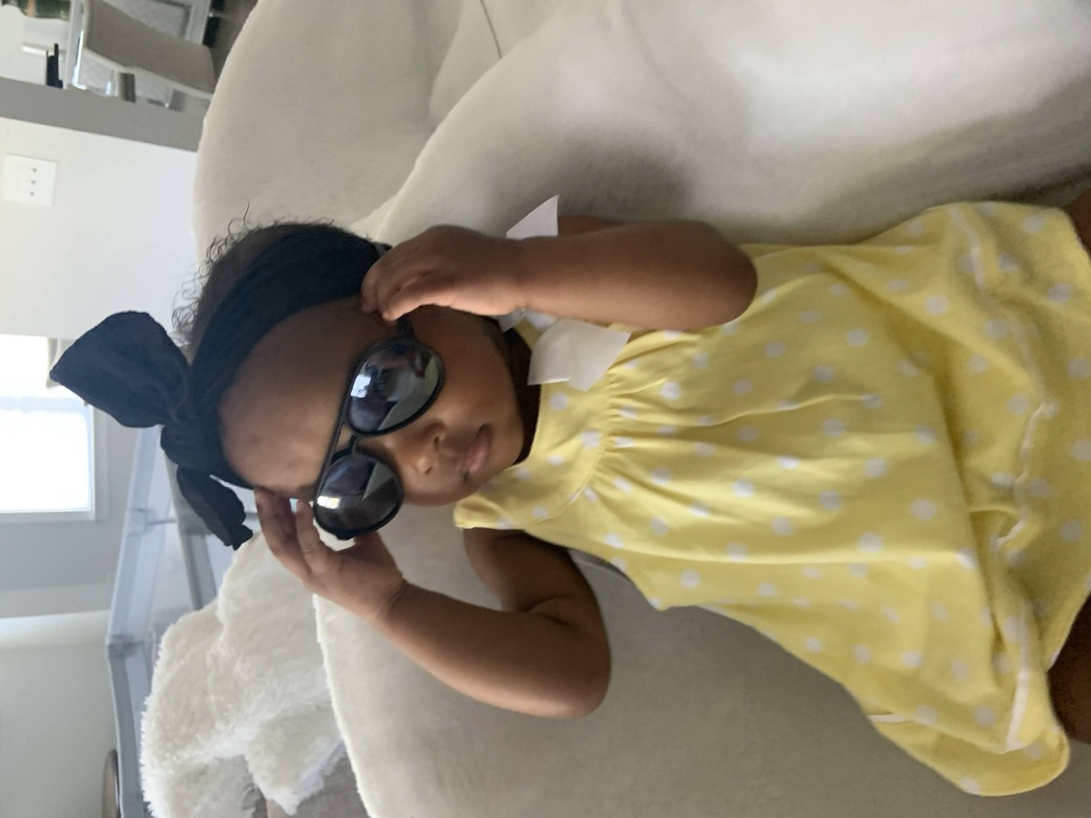 Toddler Yohanna in a yellow polka-dot dress and black bow headband, posing with oversized sunglasses on a cream chair