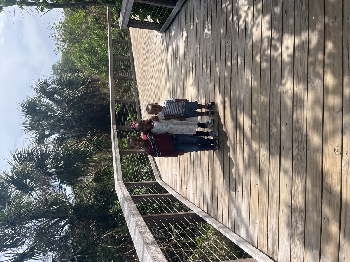 Three kids standing together on a wooden nature boardwalk surrounded by palm trees — Yohanna in a printed dress with a pink bow, flanked by her brothers