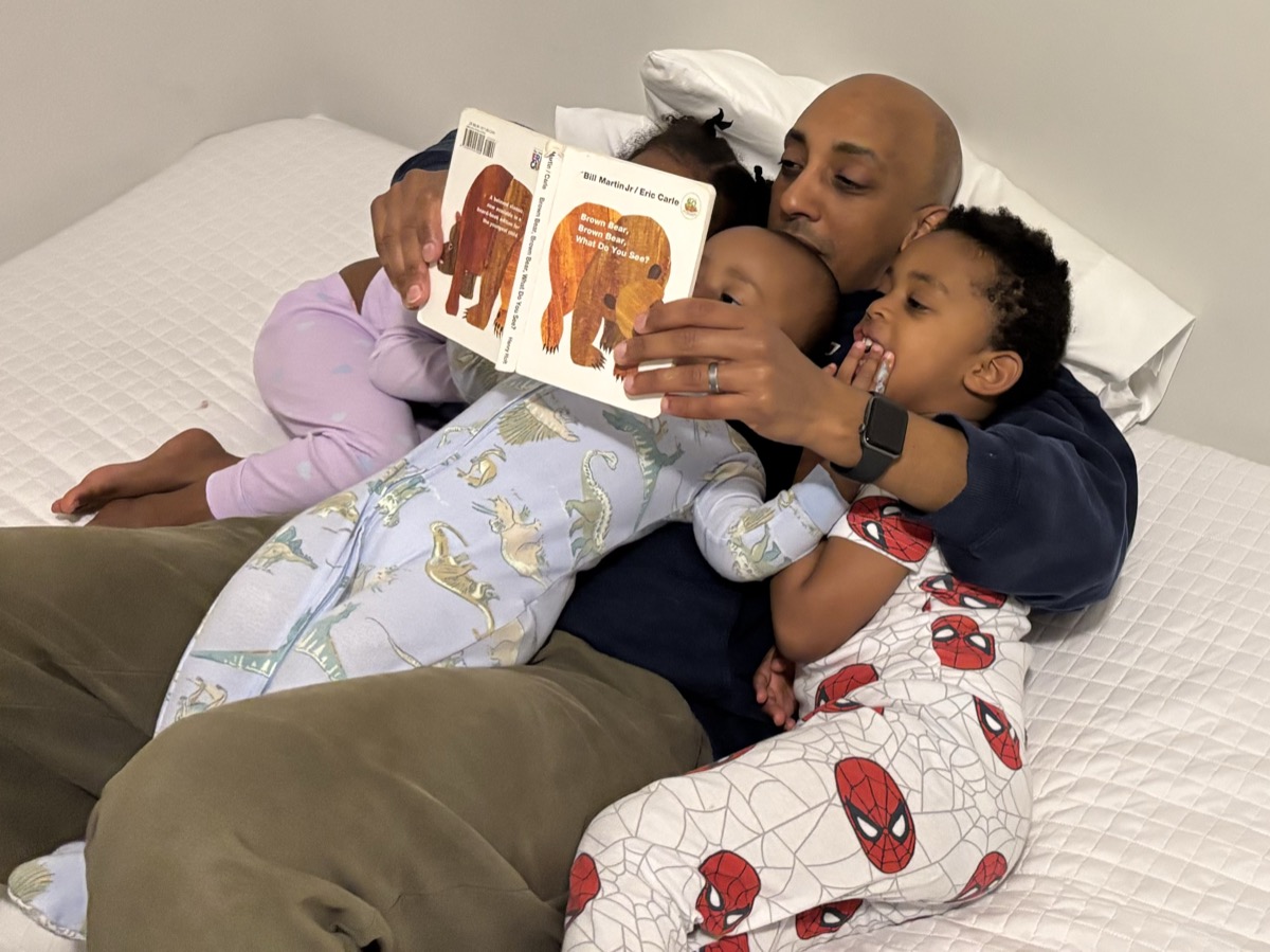Bedtime story scene — dad reading Brown Bear Brown Bear to the kids on the bed, Yohanna in dinosaur pajamas snuggled close, her brother in Spider-Man pajamas