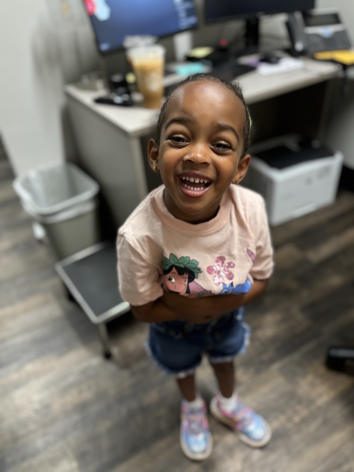 Yohanna laughing big in a pink Lilo and Stitch t-shirt and denim shorts, standing in an office at mom's workplace