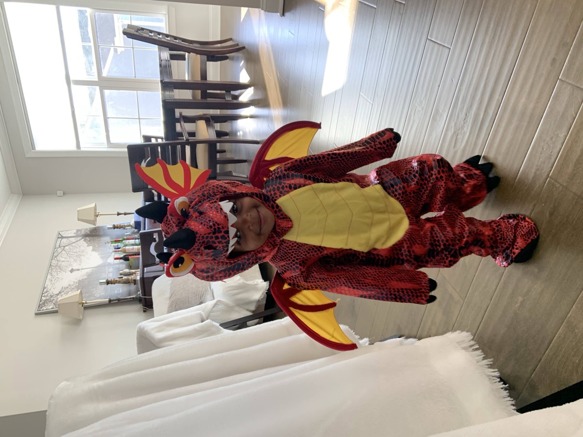 Yohanna dressed in a full red and yellow fire-breathing dragon Halloween costume, smiling in the living room