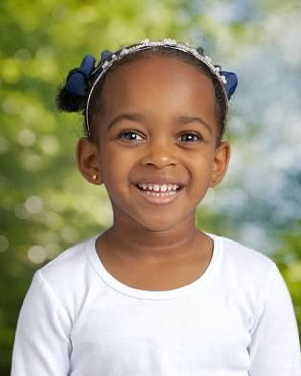 Yohanna's school portrait — beaming smile in a white top with navy bows and a sparkle headband against a soft green bokeh background