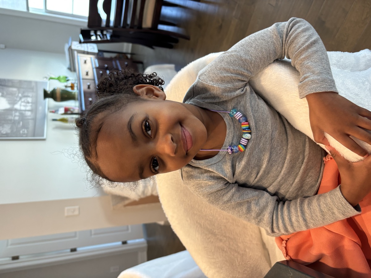 Yohanna sitting in a fluffy white chair wearing a gray top and a handmade colorful beaded necklace, smiling softly with curly hair