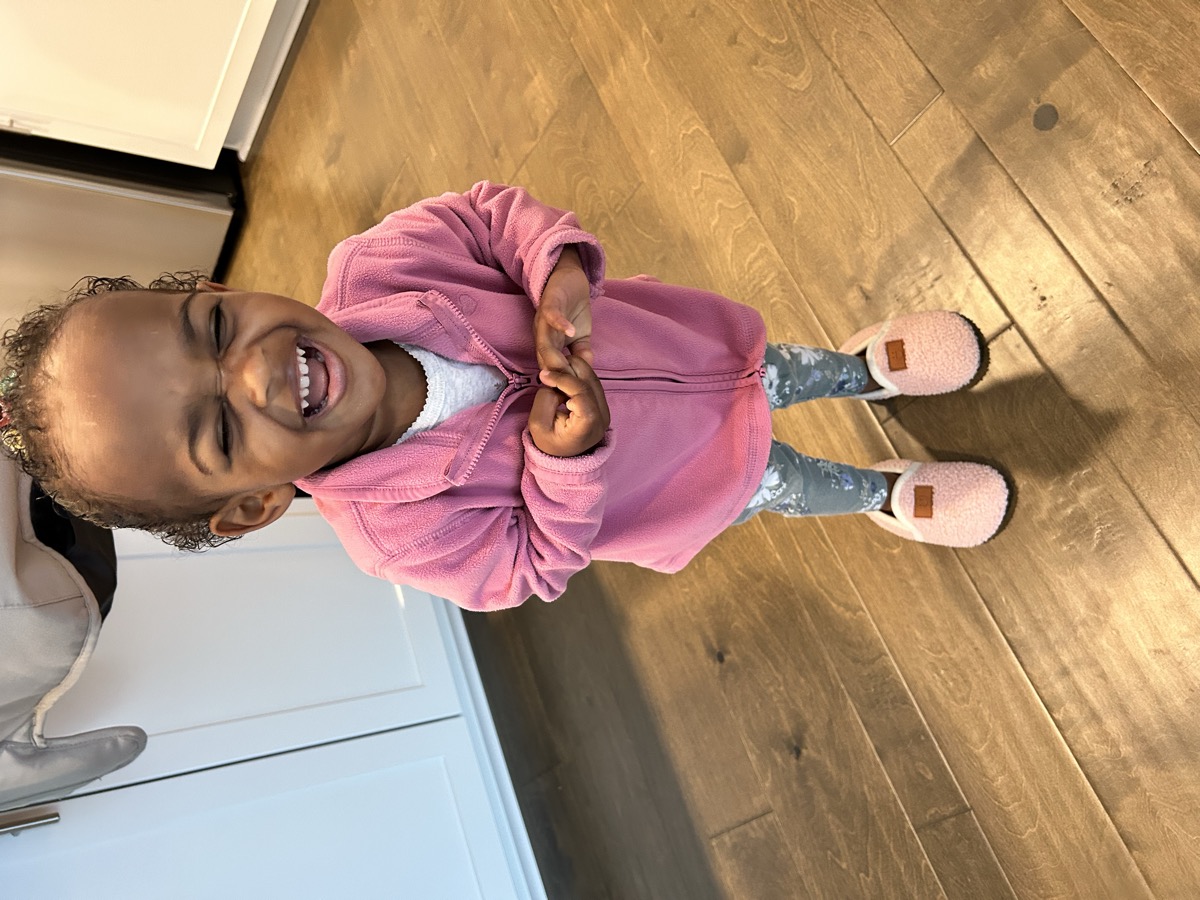 Toddler Yohanna laughing hard in a pink fleece jacket, floral leggings, and fuzzy pink slippers on hardwood floor