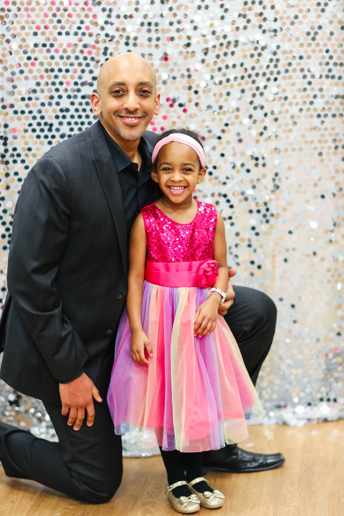 Yohanna and dad at a party — she is in a sparkly pink sequin dress with a rainbow tulle skirt and pink headband, he is in a dark suit, both smiling in front of a sequin backdrop