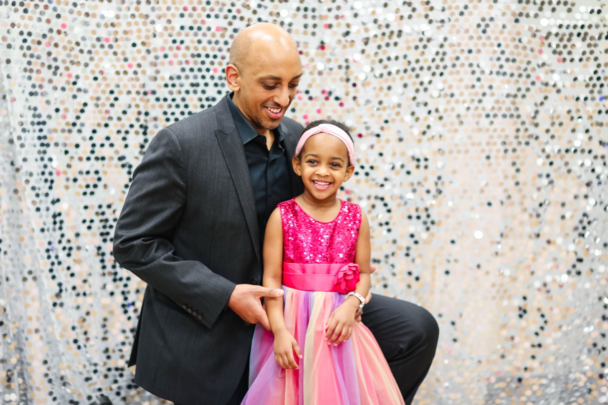 Dad looking lovingly at Yohanna in her pink sequin party dress in front of a glittery silver backdrop