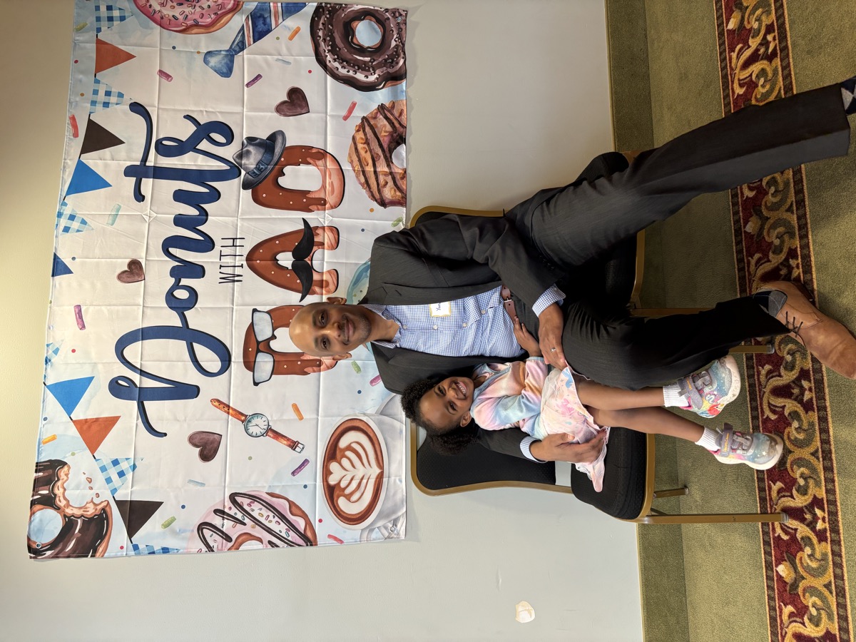 Yohanna sitting on dad's lap at a Donuts with Dad event, wearing a pastel tie-dye jacket and smiling