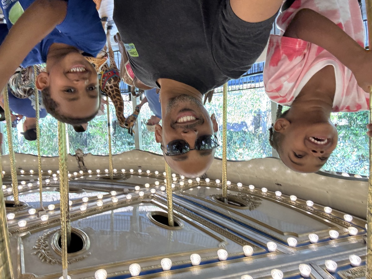 Yohanna, dad, and her twin brother on a carousel — Yohanna in a pink tie-dye shirt looking up with joy, dad in sunglasses, brother in a blue shirt