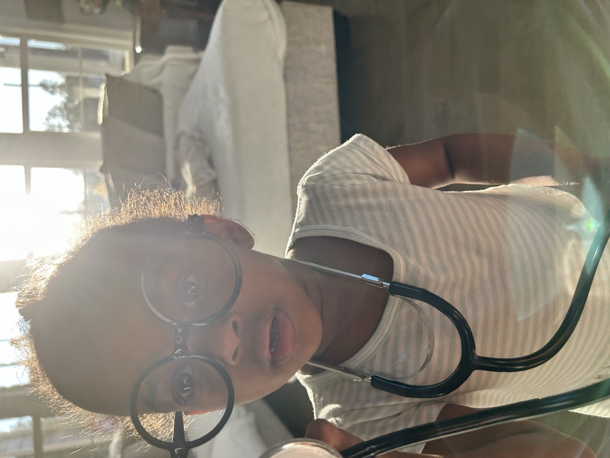 Toddler Yohanna wearing oversized round glasses and a stethoscope in a striped onesie, playing doctor with golden backlight