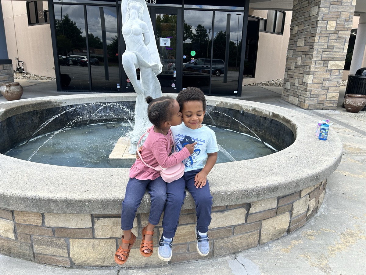 Yohanna in a pink jacket and crossbody bag giving her brother a kiss on the cheek while sitting on the edge of an outdoor fountain