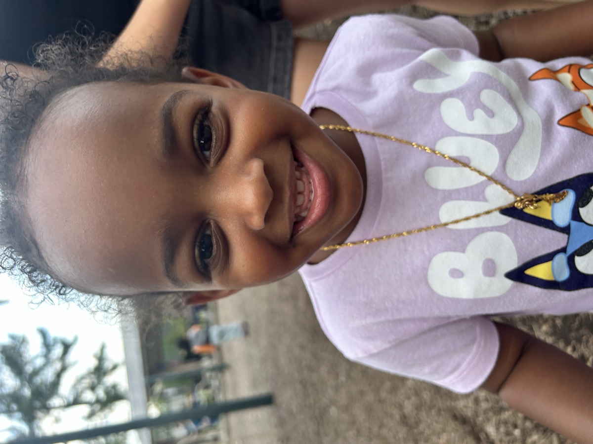 Close-up of Yohanna at the playground in a pink Bluey t-shirt with a gold chain necklace, smiling big