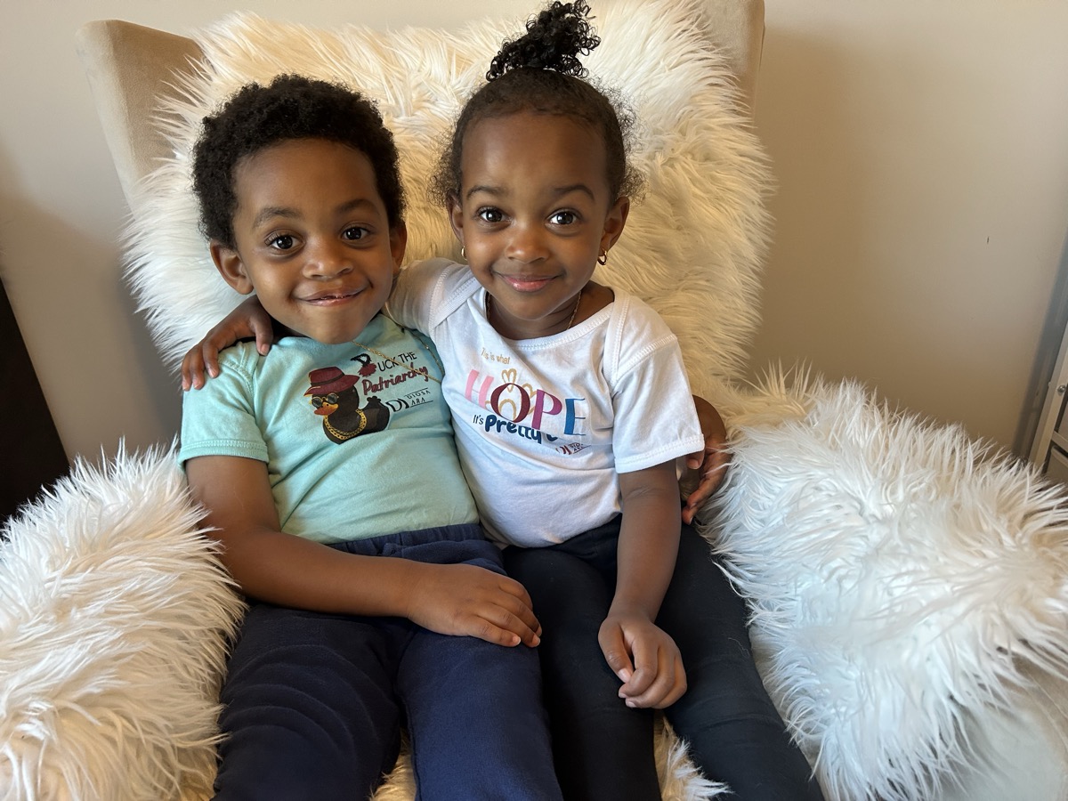 Yohanna and her twin brother sitting together on a fluffy white chair — she in a HOPE t-shirt with a ponytail, he in a green graphic tee, arms around each other