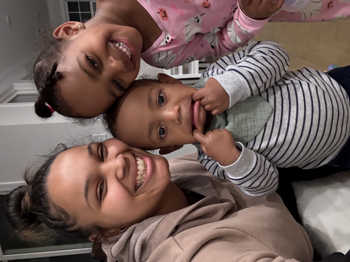 Yohanna in pink Halloween pajamas leaning into a selfie with a young woman in a beige hoodie and a toddler boy in a striped shirt making a silly face