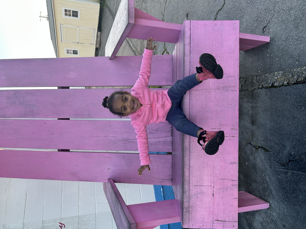 Yohanna sitting in an oversized pink Adirondack chair with arms stretched wide, wearing a pink hoodie, jeans, and sparkly pink boots