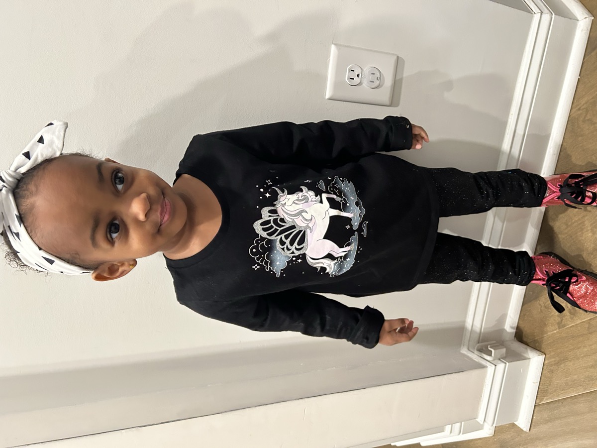 Yohanna standing indoors in a black unicorn sweatshirt, sparkle black leggings, pink glitter boots, and a black-and-white patterned headband bow