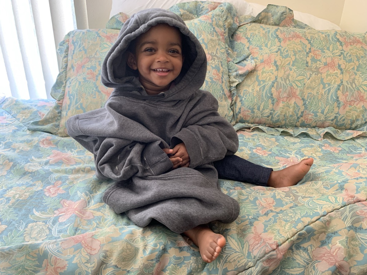 Yohanna sitting on a floral bedspread in an oversized gray hoodie with the hood up, smiling happily