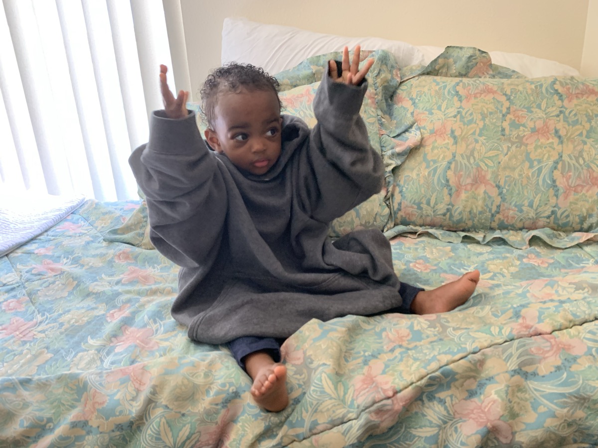 Yohanna on the floral bed in an oversized gray hoodie with arms raised playfully, looking to the side