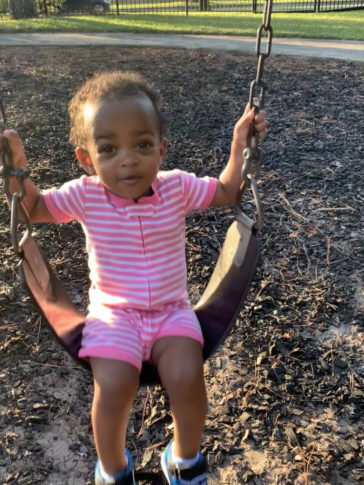 Baby Yohanna on a playground swing in a pink striped romper, holding the chains and looking at the camera