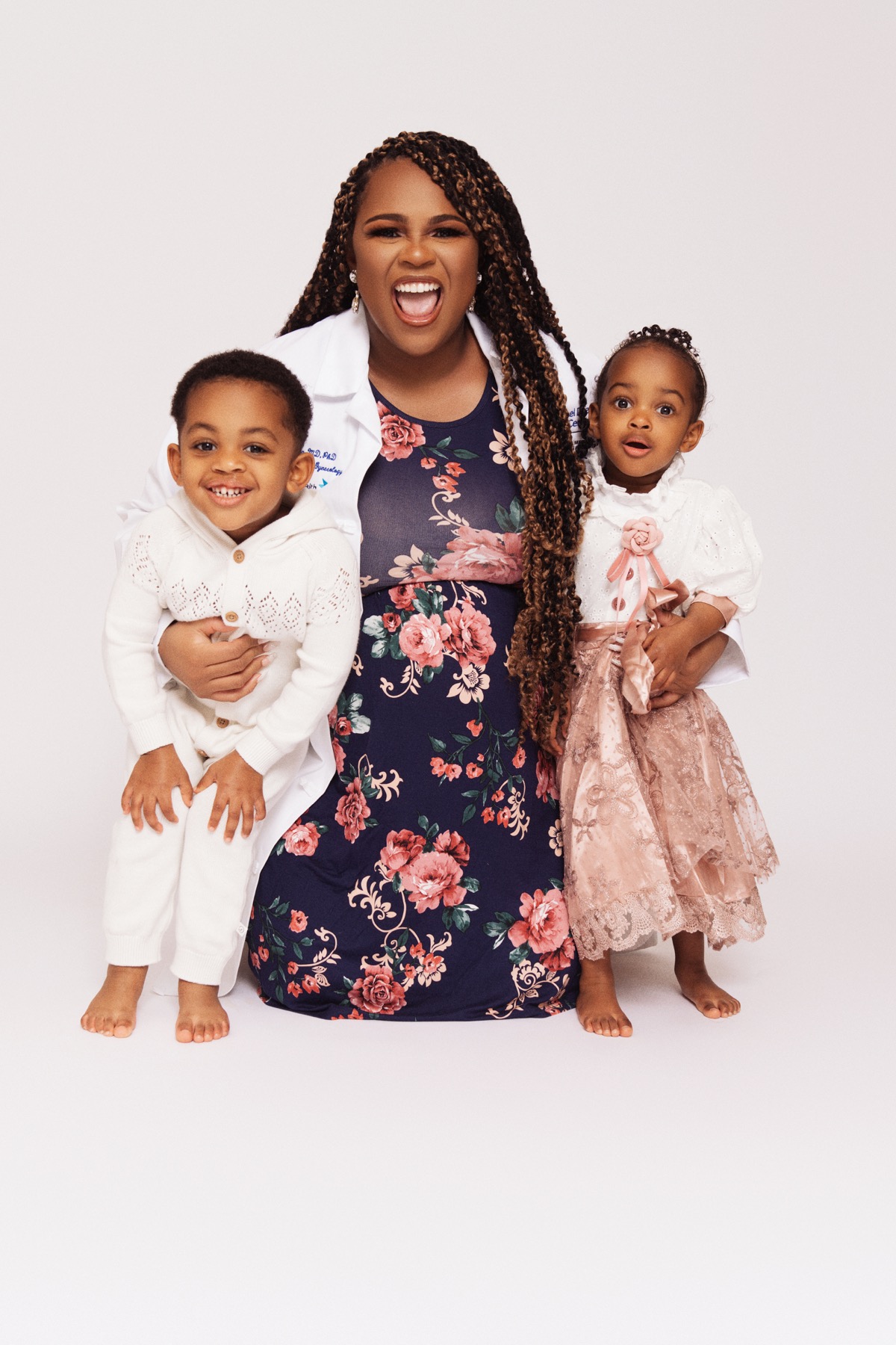 Professional studio portrait — mom in a white doctor's coat and floral dress holding both children, Yohanna in a pink lace dress with a matching bow, her brother in a white outfit